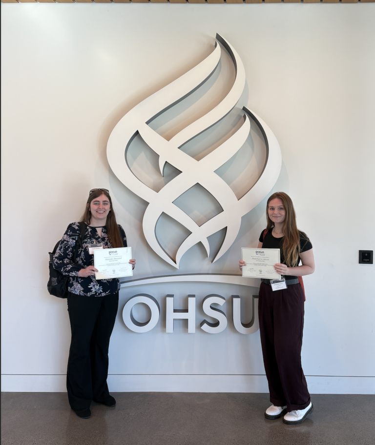Hannah and Anna with their travel awards in front of OHSU logo